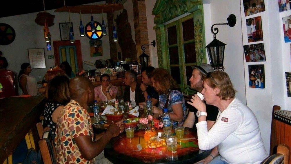 A group of people seated around a table with drinks and snacks, in a room with a green window and wall decorations. 4
