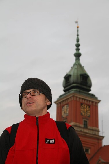 A person wearing a red vest with a logo, standing in front of a clock tower with a green spire. 1