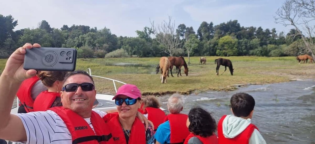A group of people on a boat, wearing life jackets, with horses grazing on the shore. 1