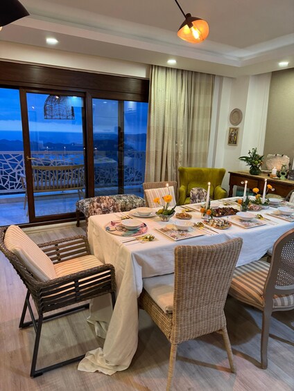 A dining room with a table set for a meal, wicker chairs, a green armchair, and a view of the ocean through large windows. 3