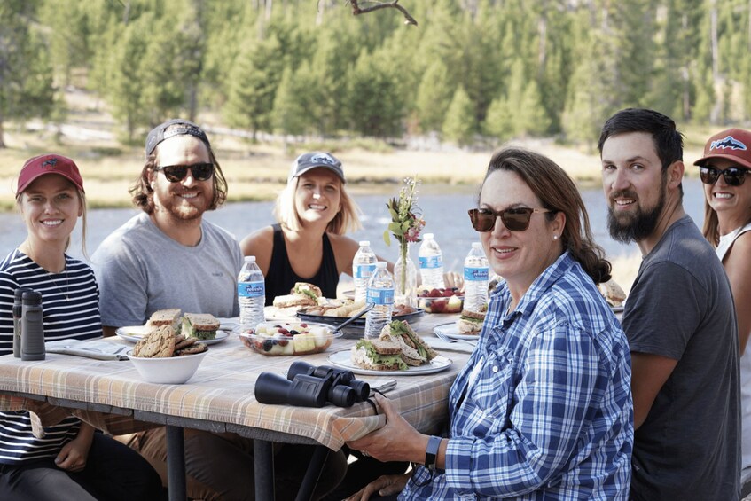 A group of people sitting at a picnic table with food and drinks, surrounded by a forested area. 3
