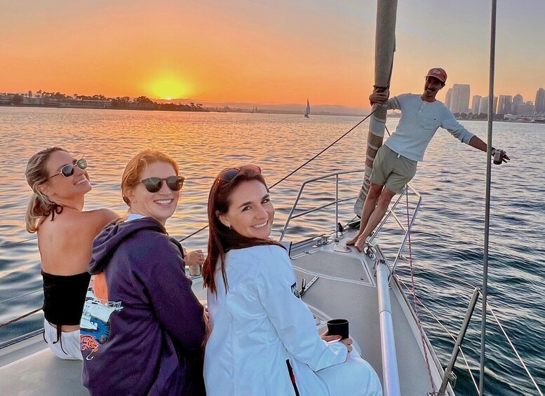 A sunset on a calm body of water with a sailboat, a city skyline in the background, and three people on the deck. 1