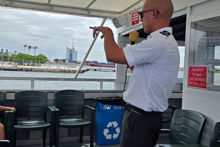 A man in a white shirt and dark pants standing on a boat, pointing towards the horizon. There are green chairs and a blue recycling bin on the deck. A sign indicates a limit of 149 adults and 15 children on the first deck. 5
