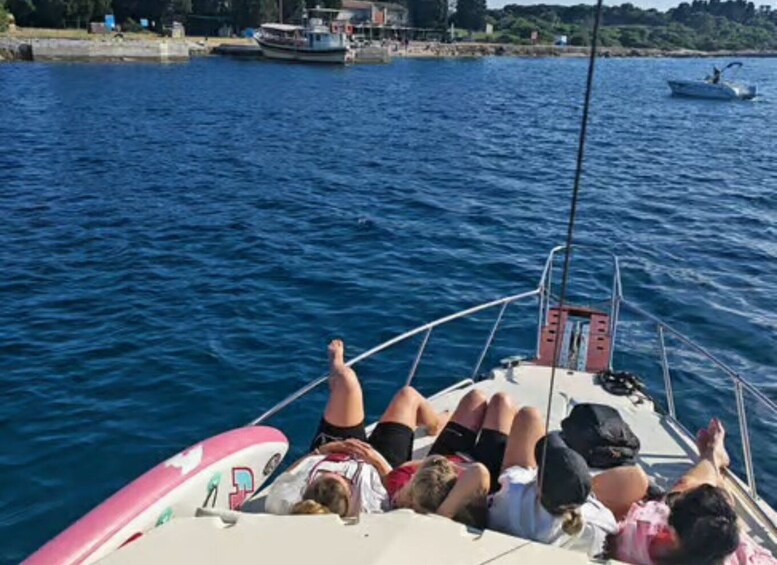 A boat with people lying on deck, calm water, and a shoreline with buildings and trees in the background. 1