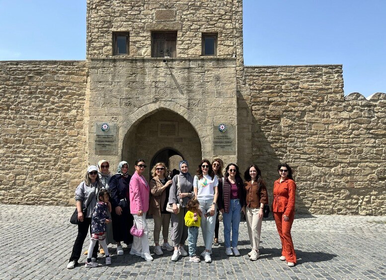 A group of people standing in front of a stone building with an arched entrance and a tower. 1