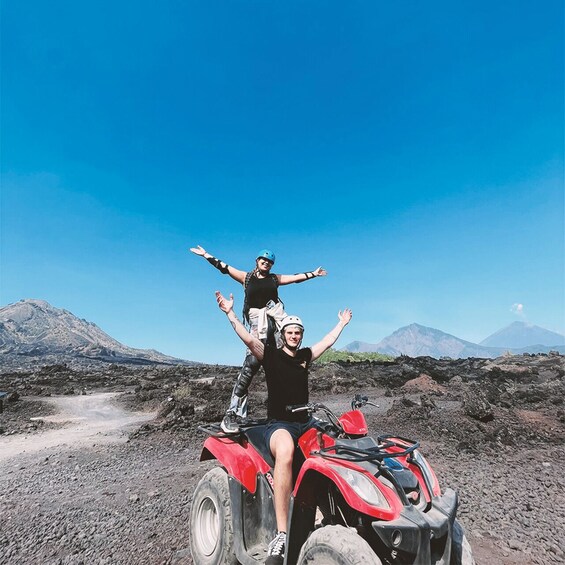Two people on a red ATV with a rocky landscape and mountains in the background. 1