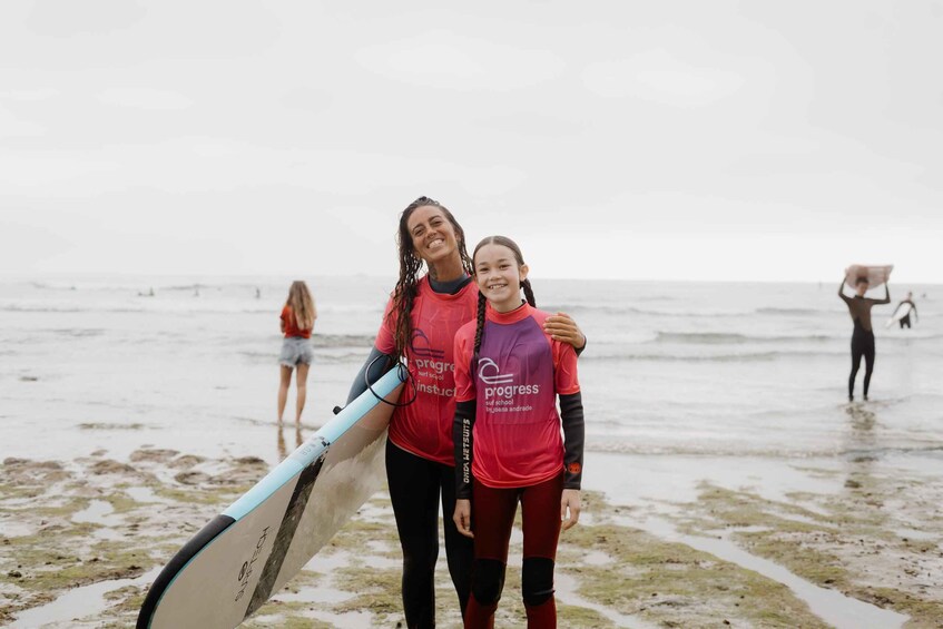 Two people wearing matching red and purple wetsuits with 'progress' text, holding a surfboard on a beach with other people in the background. 5