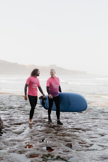 Two people in pink shirts walking on a rocky beach with a blue surfboard. 4