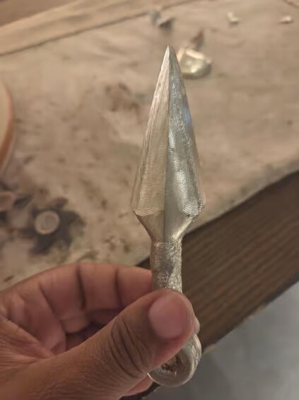 A hand holding a metallic object with a pointed tip, possibly a tool or weapon, against a cluttered background with a wooden surface. 4