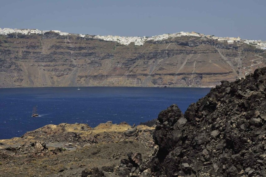 A coastal landscape with white buildings on a cliff, a rocky foreground, and a sailboat on the water. 1