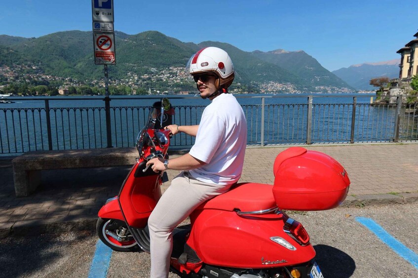 A person on a red scooter with a white helmet, parked by a lake with mountains in the background. 1