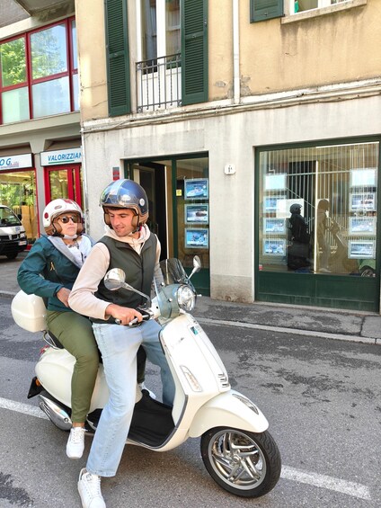 A white scooter with two riders, one in a green jacket and the other in a beige jacket, parked in front of a building with green shutters and a sign that reads 'VALORIZZIANI'. 2