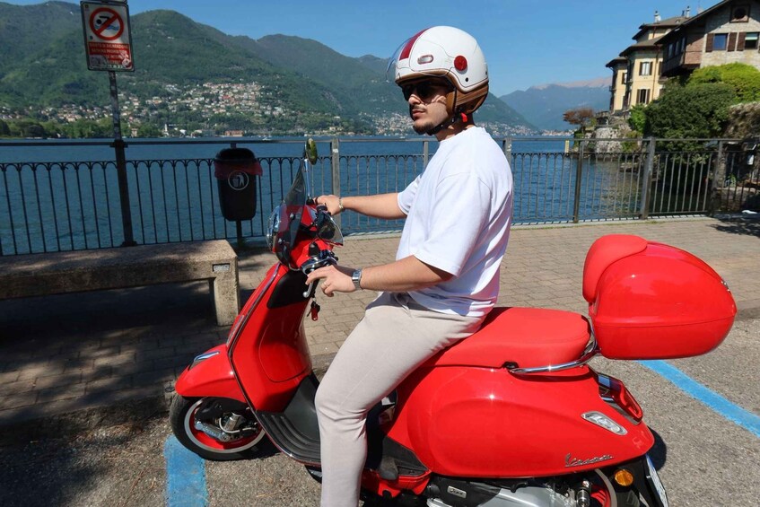 A person on a red scooter with a helmet, near a waterfront with mountains in the background. 4
