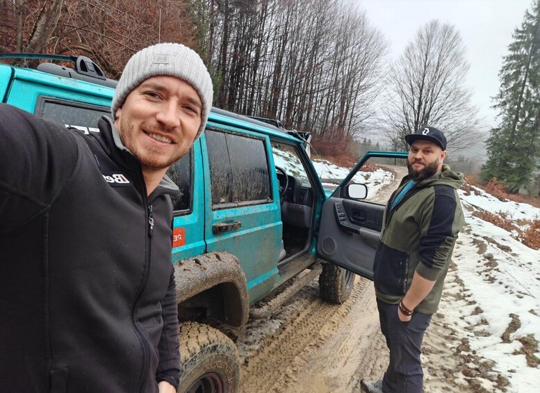 Two people near a blue off-road vehicle on a snowy path with trees in the background. 4