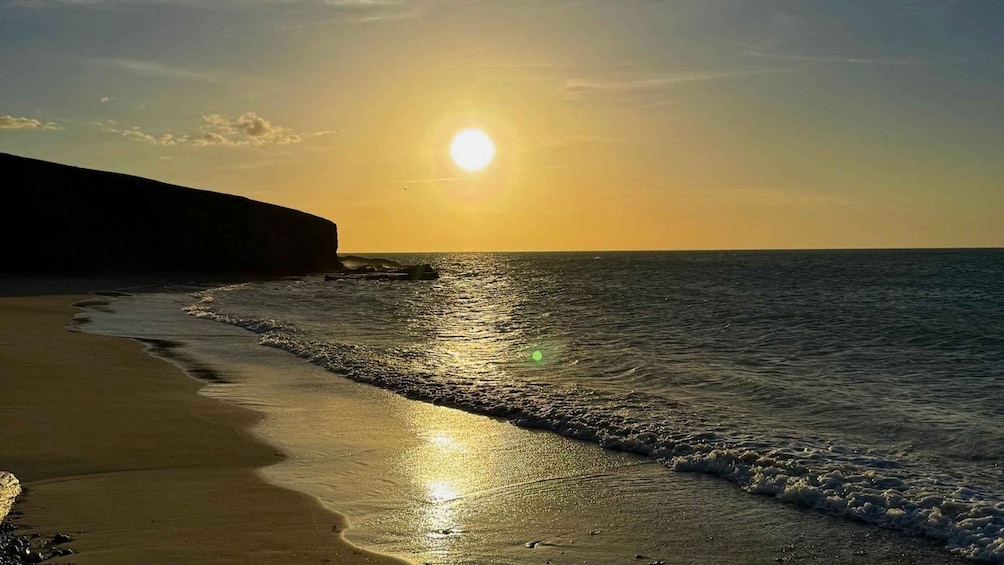 A sunset over a beach with waves lapping at the shore and a cliff in the background. 1