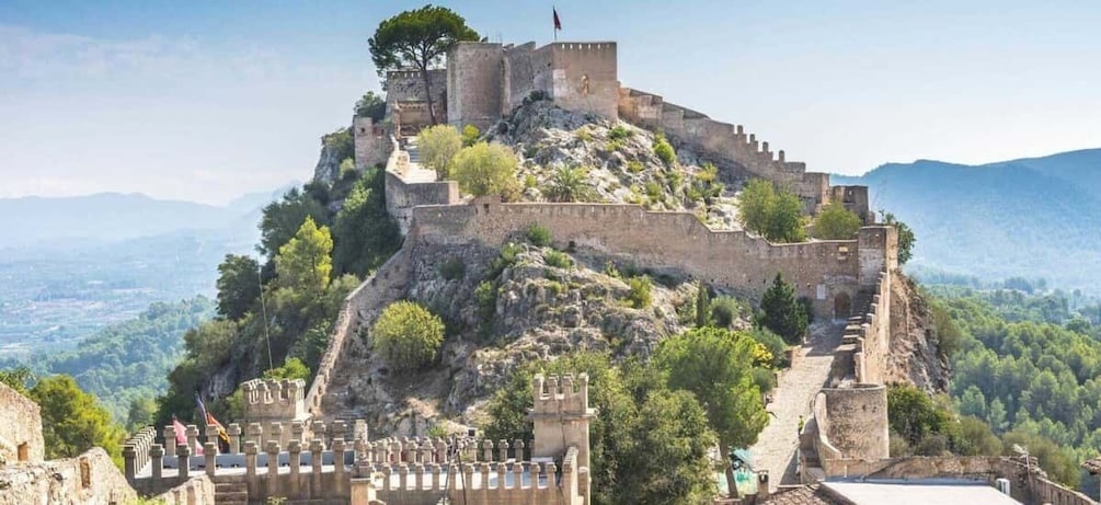 A historic stone fortress on a hill with a flag, surrounded by greenery and mountains. 4
