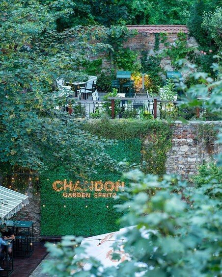 A garden with a neon sign reading 'CHANDON GARDEN SPRITZ' amidst lush greenery and stone walls. 1