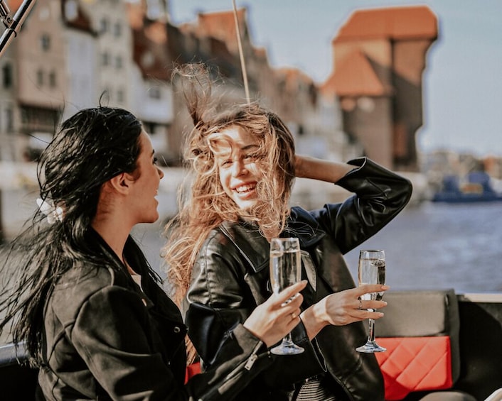 Two people on a boat, holding champagne glasses, with a cityscape in the background. 1