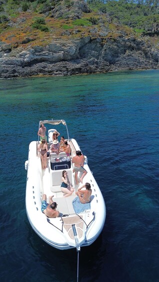 A small inflatable boat with people on it, anchored in clear blue water with rocky terrain and greenery in the background. 1