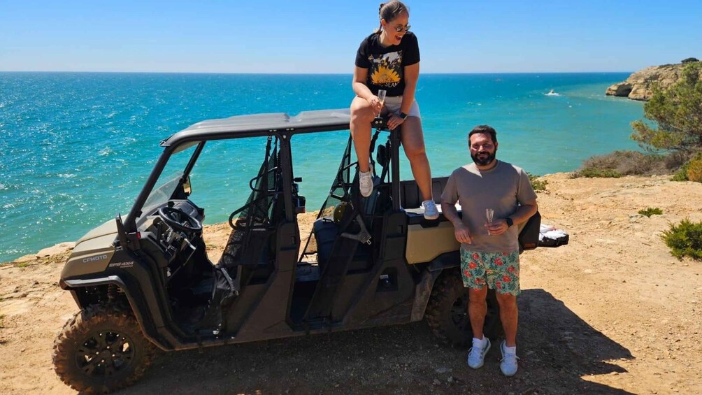 A man and a woman standing next to a golf cart by a coastal area with clear blue water and rocky terrain. 1