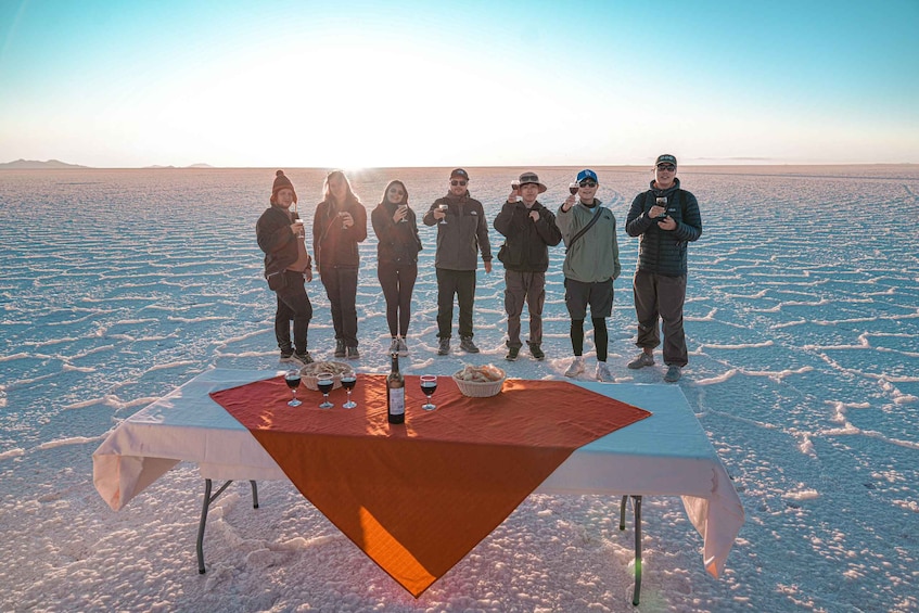 A group of people standing around a table with a bottle and glasses on it in a snowy landscape. 2