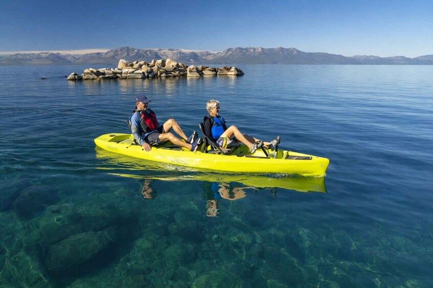 Two people in a yellow kayak on a calm lake with clear water, surrounded by mountains and rocks. 3