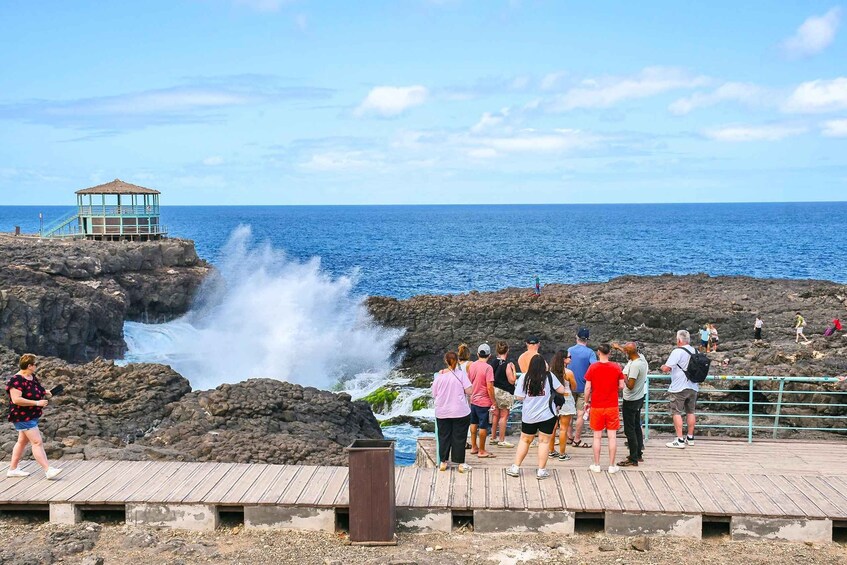 A rocky coastline with a wooden walkway, a gazebo, and people observing a waterfall. 2