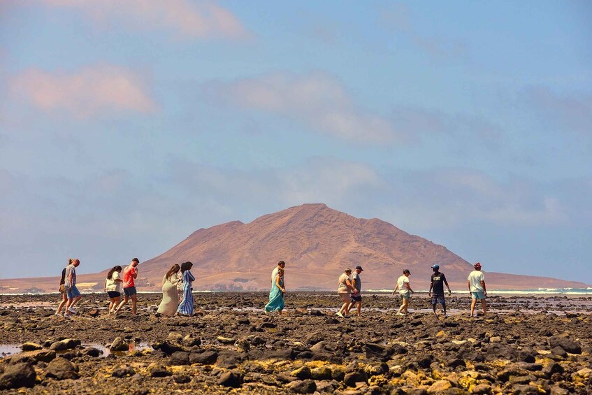 A group of people walking on a rocky beach with a mountain in the background. 1