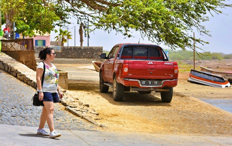A woman walking on a cobblestone path, a red pickup truck parked on a sandy area, and boats in the water. 5