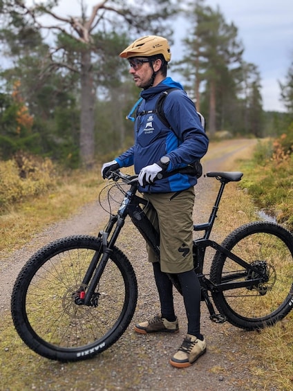 A person standing next to a mountain bike on a gravel path with trees in the background. 1
