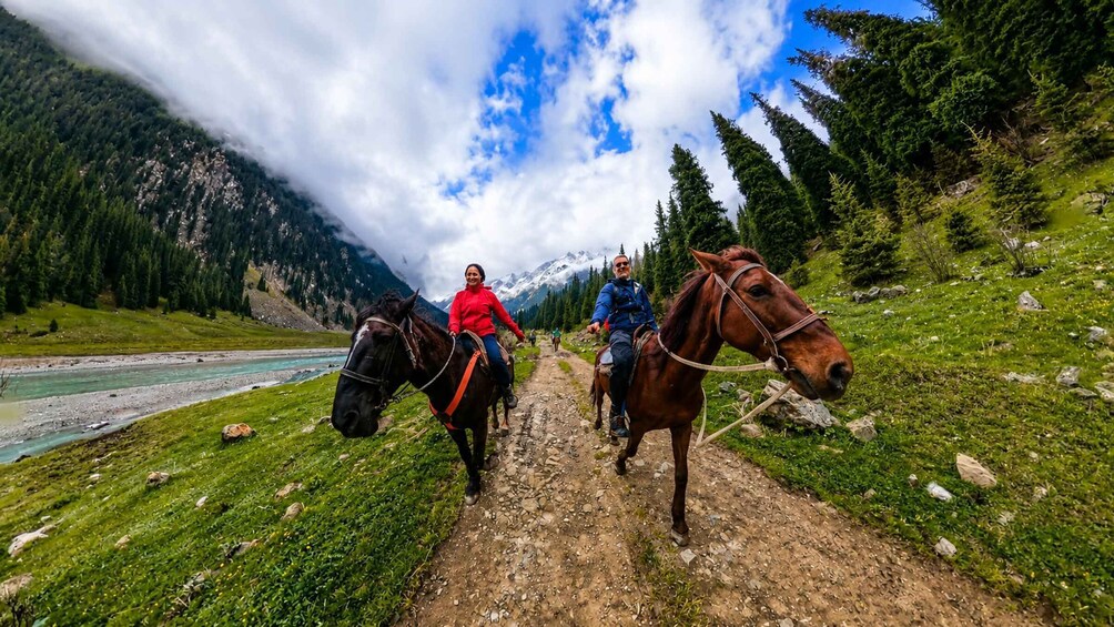 A group of people horseback riding on a trail with a river and mountains in the background. 4
