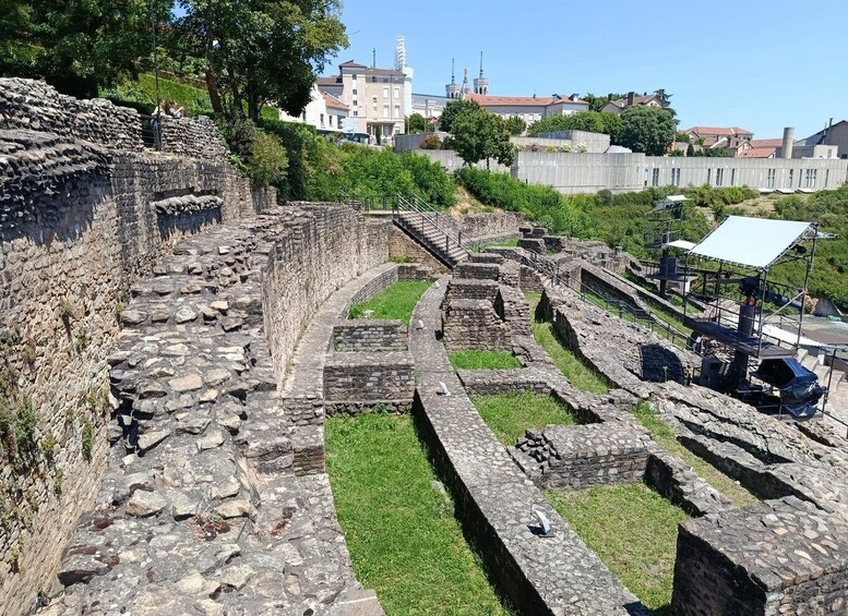 Ancient stone ruins with a grassy pathway and modern buildings in the background. 3