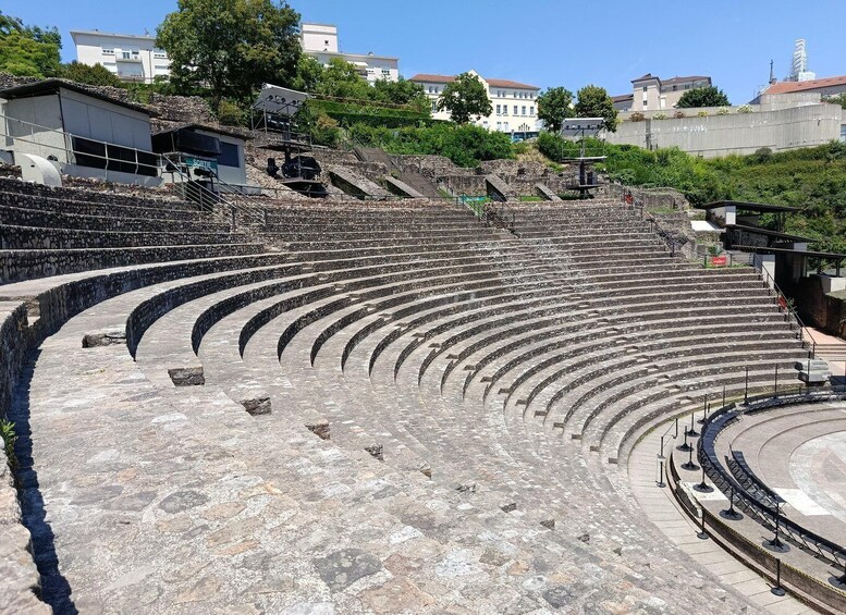 An ancient amphitheater with stone steps and a central stage, surrounded by modern buildings. 1