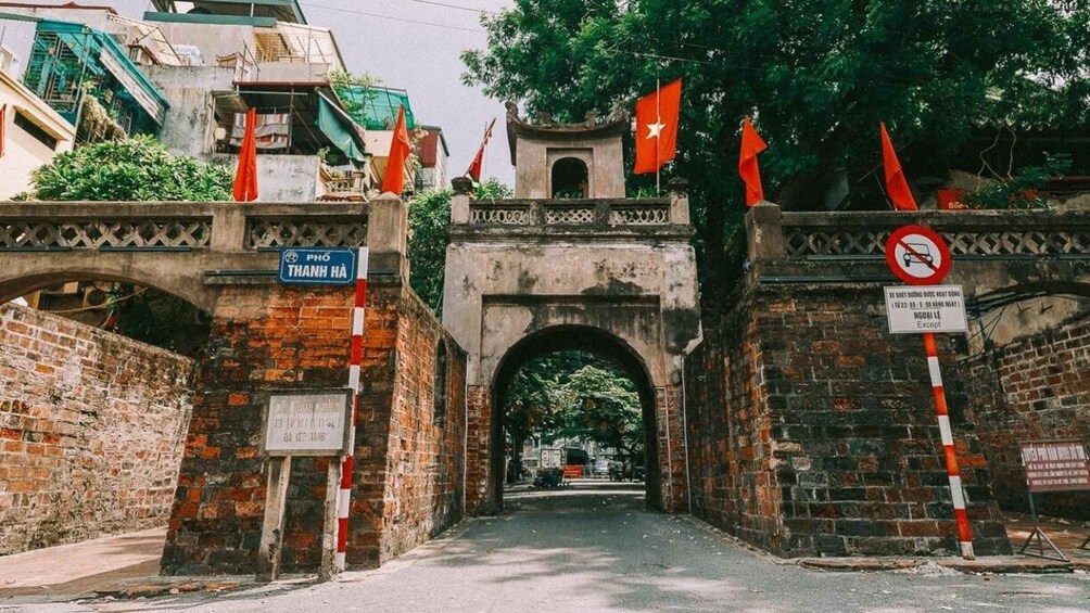 A historic stone archway with red flags, a no parking sign, and a sign reading 'PHÒ PHÒ THÀNH HÀ'. 1