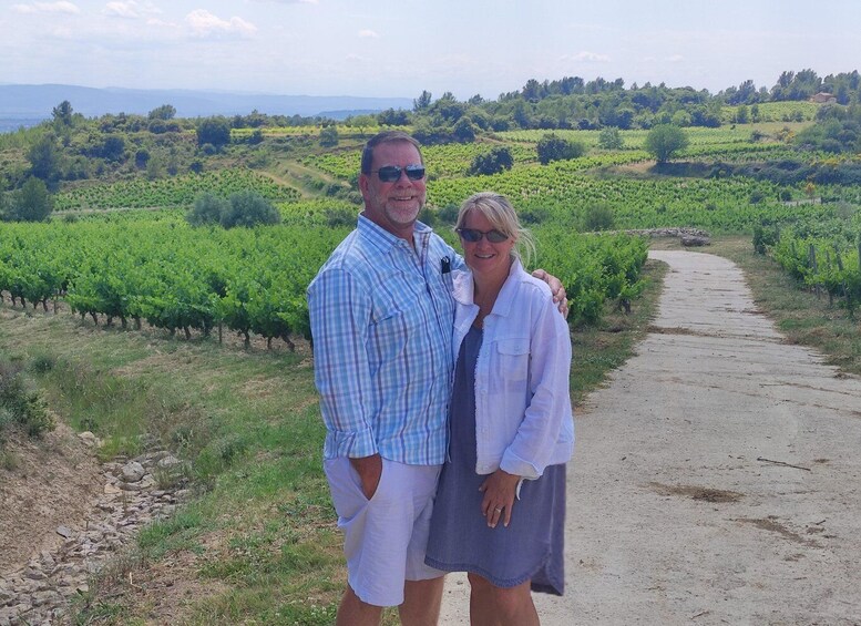 A man and woman standing on a dirt path with vineyards in the background. 1