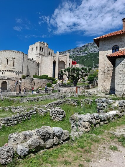 A historic stone building with a red flag, a mountainous backdrop, and a clear blue sky. 3