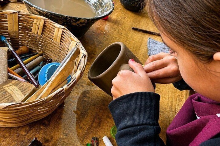 A person crafting pottery on a wooden table with a basket of tools and a clay pot. 1