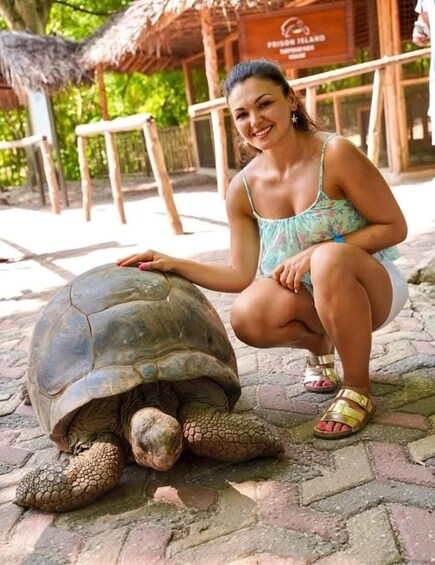 A person crouching next to a large tortoise on a paved area with a thatched-roof structure in the background. 5