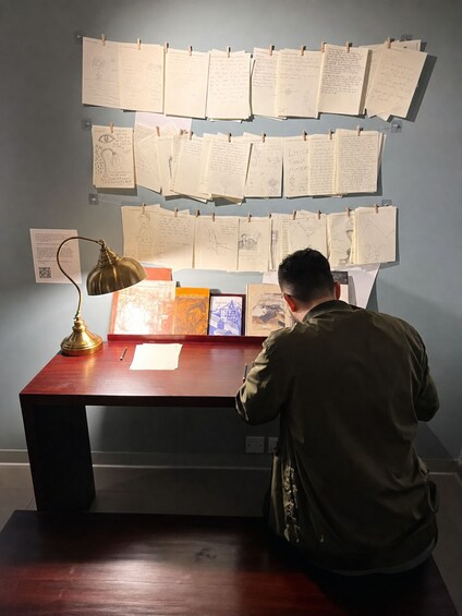 A person at a desk with papers pinned to the wall, a lamp, and a framed picture. 4
