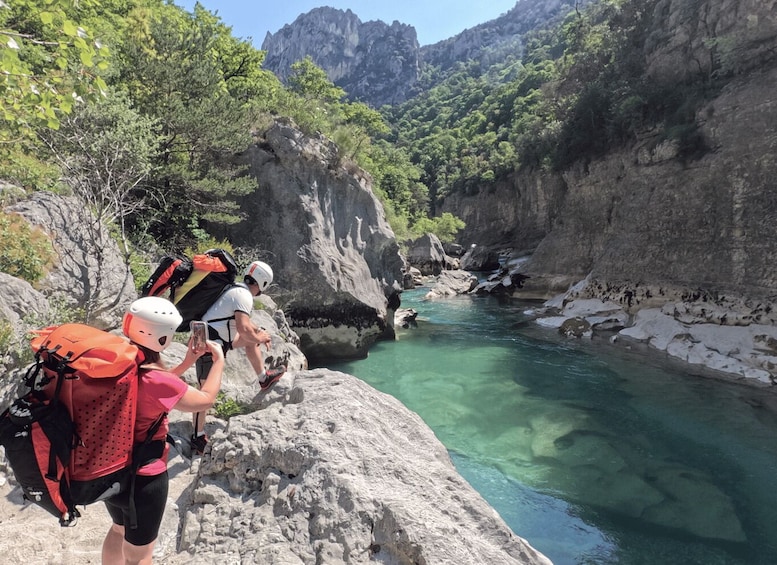 Hikers on a rocky riverbank with clear water and mountainous terrain in the background. 3