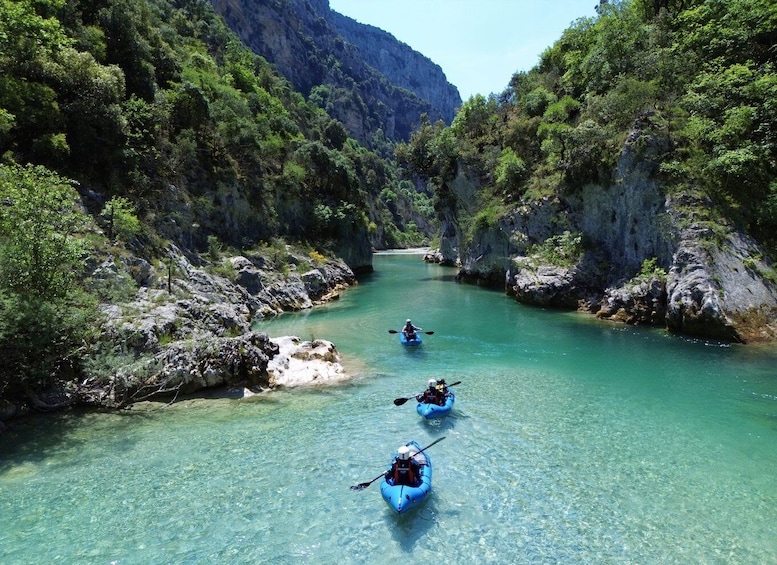 Three kayakers on a clear river surrounded by rocky cliffs and dense greenery. 1