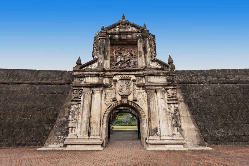 Un portón de piedra ornamentado con intrincados grabados y un emblema central, que da paso a un camino de ladrillo. 1