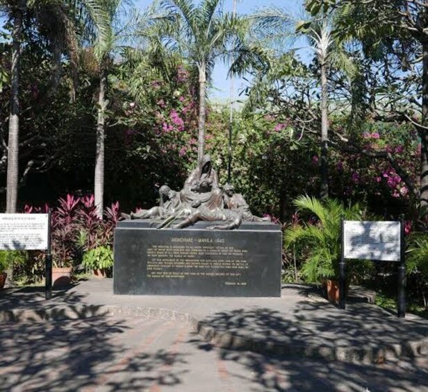 Una estatua de una figura histórica rodeada de vegetación tropical y dos placas informativas. 4