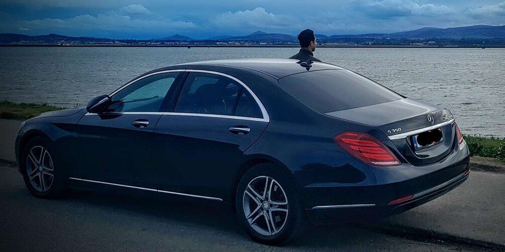 A dark-colored sedan parked by a waterfront with a person standing on the roof. 1