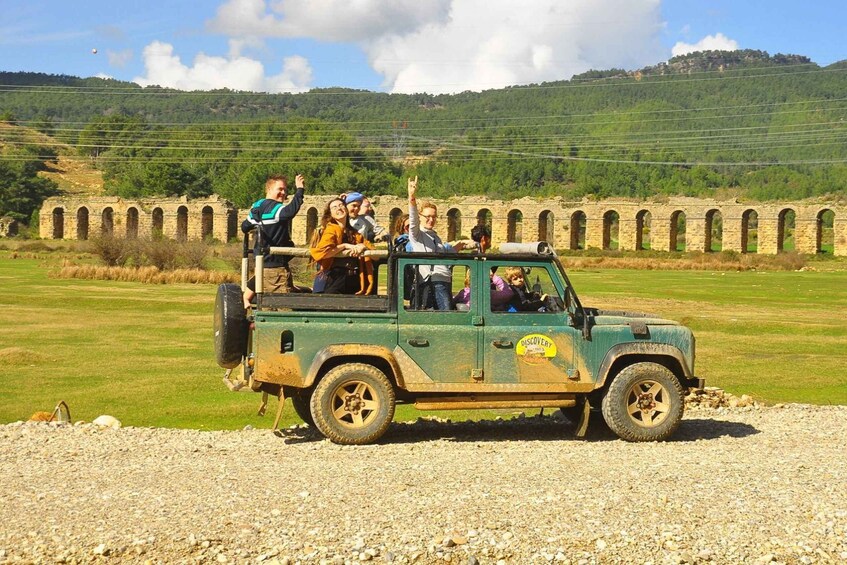 A green off-road vehicle with passengers, parked on a gravel area with a stone viaduct and green hills in the background. 5