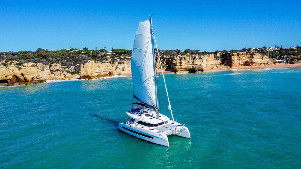 A sailboat on turquoise waters with a rocky coastline and buildings in the background. 3