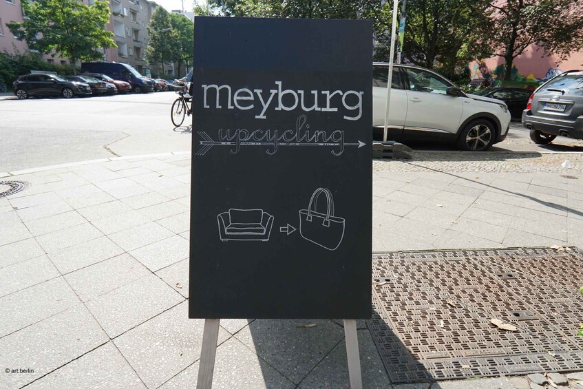 A signboard on a sidewalk advertising 'meyburg upcycling' with illustrations of a sofa and a handbag. 5