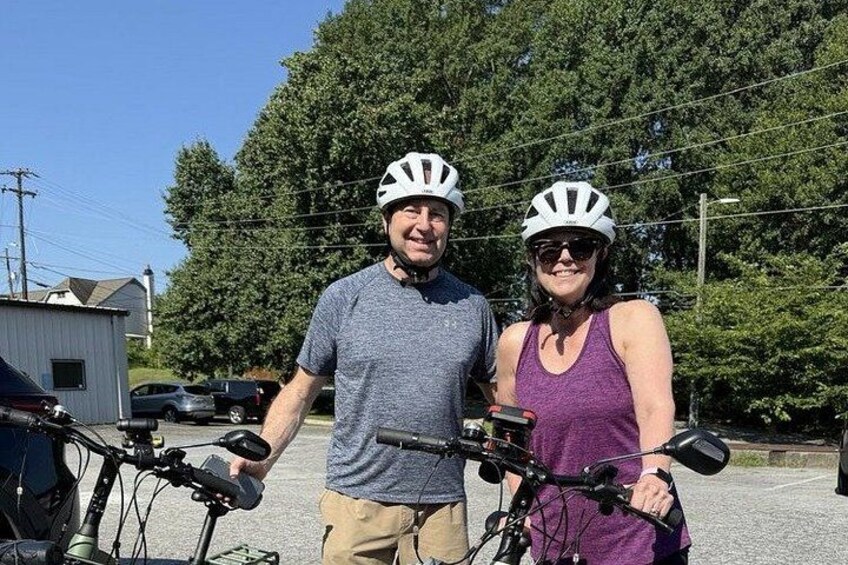 Two people with bicycles, wearing helmets, in a parking lot with trees and a building in the background. 3