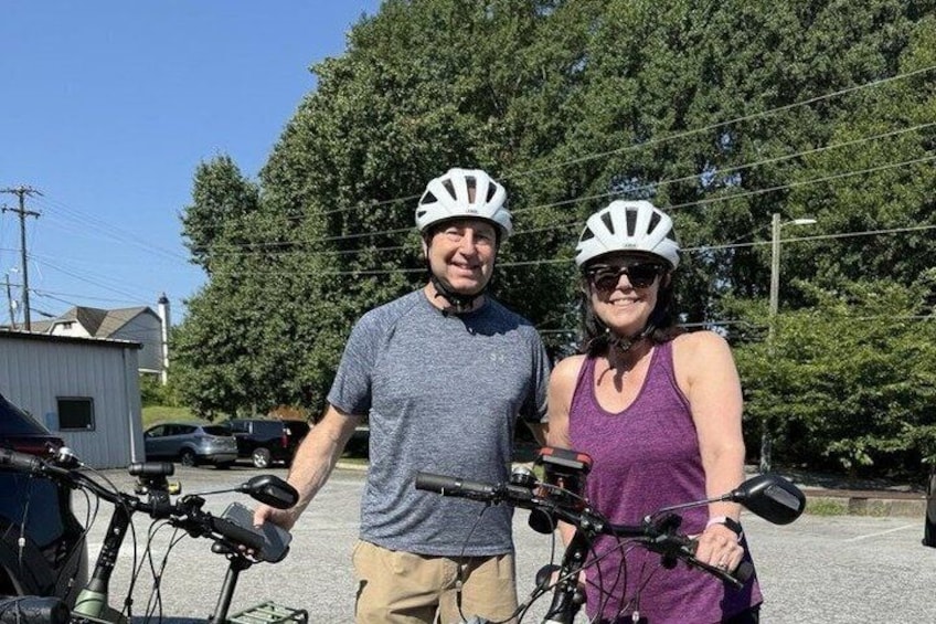 Two people with bicycles and helmets, standing in a parking lot with trees and a building in the background. 5