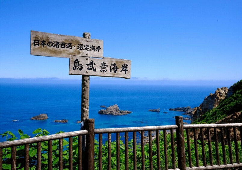 A wooden signpost with Japanese text, overlooking a coastal landscape with clear blue sea and rocky outcrops. 5